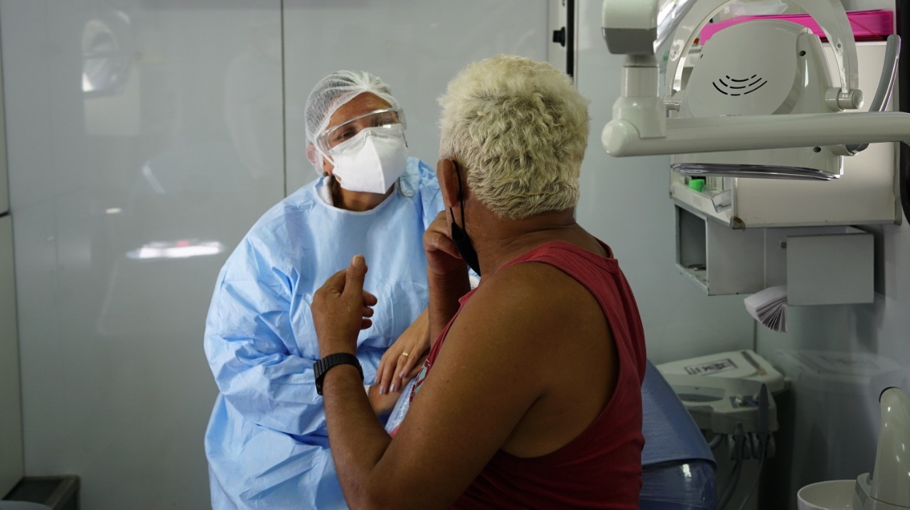 Na imagem há uma profissional da saúde sentada em uma cadeira enquanto olha para o paciente que está sentado em sua frente de costas para a foto e parece estar mostrando a boca para a dentista. O paciente tem o cabelo loiro e usa uma regata cor vermelho escuro. 